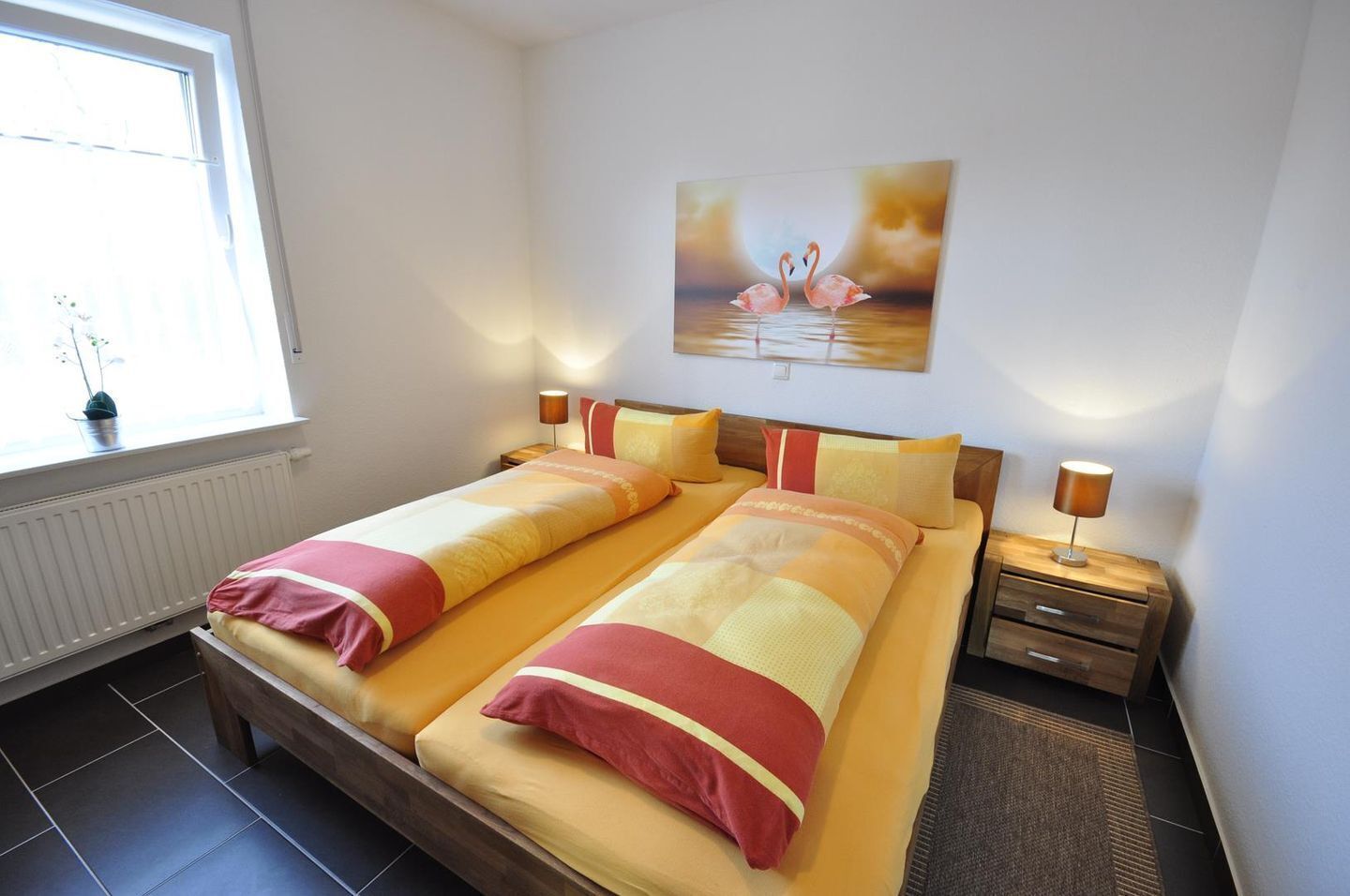 Ein Doppelbett mit passenden Kissen, ein Nachttisch mit Lampe, ein Fenster mit Blick auf Grünpflanzen und ein Flamingo-Gemälde an der Wand.