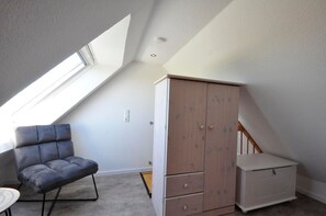 Una habitación con techo inclinado, un sillón tapizado en gris, un armario de madera y una pequeña mesa blanca.