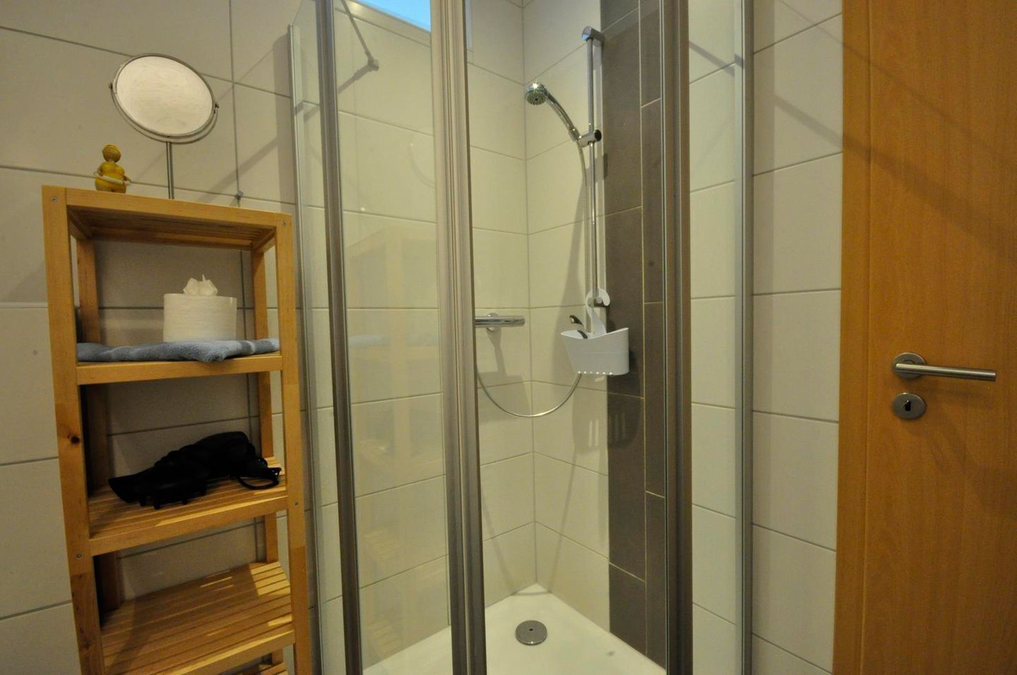 A shower area with a glass door, a wooden shelf with towels and a black item, and a white trash bin.