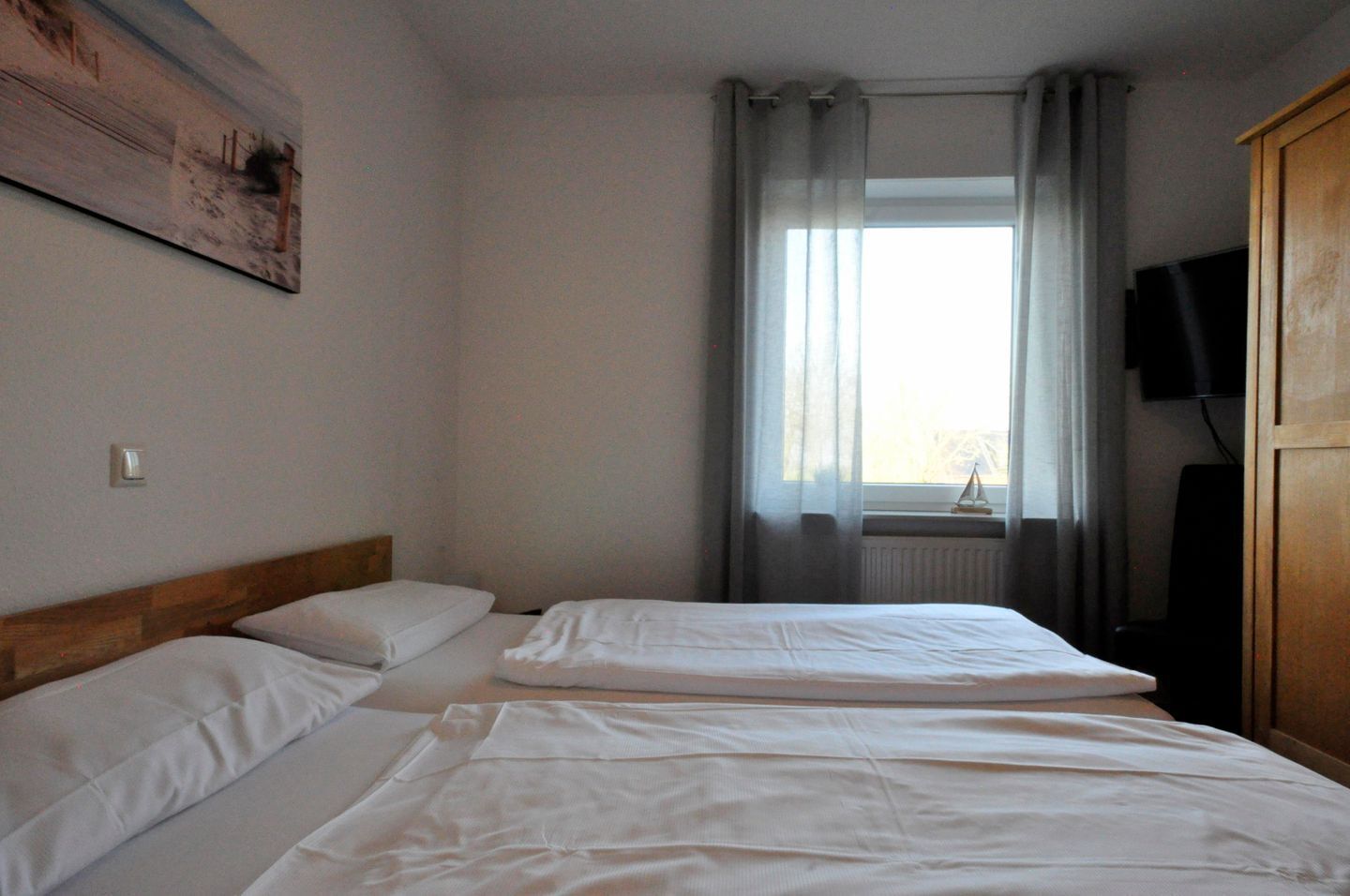 Una habitación con dos camas, una ventana con cortinas, un televisor montado en la pared y un armario de madera.