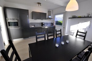 Une cuisine moderne avec des meubles de couleur sombre, un four encastré et un coin repas comprenant une table et des chaises noires.