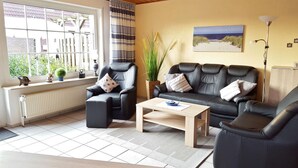 Ein Wohnzimmer mit einem schwarzen Ledersofa, einem schwarzen Leder-Sessel und einem Couchtisch aus Holz. Ein Fenster bietet Blick auf Grünflächen.