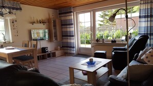 Ein gemütliches Wohnzimmer mit Holzbalkendecke, einem Essbereich mit Tisch und Stühlen, einer schwarzen Ledersofa und einem großen Fenster mit Vorhängen.