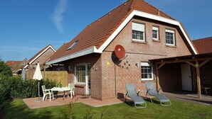 Ein zweistöckiges Ziegelhaus mit geneigtem Dach, einer überdachten Terrasse mit zwei Liegestühlen und einem eingezäunten Garten.