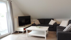 Un salon avec un téléviseur à écran plat, une table basse blanche, un canapé noir et une fenêtre avec des rideaux.