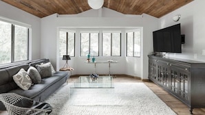 En moderne stue med træloft, en grå sofa med mønstrede puder, et glastæppe og et mørkt træskab.