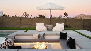 A modern outdoor seating area with a fire pit, white cushions, and an umbrella. - Ventana Cove Pickleball and Saltwater Pool (Yucca Valley)
