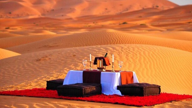 A desert dining setup with a table and cushions on a red carpet, set against a backdrop of sand dunes. - Camel trek bivouac in Merzouga (Merzouga)