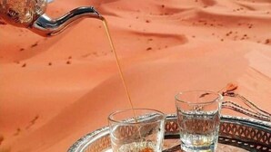 A tray with two glasses, one with a dark liquid and the other with a clear liquid, and a metal pitcher pouring a golden liquid into the clear glass. - Camel trek bivouac in Merzouga (Merzouga)
