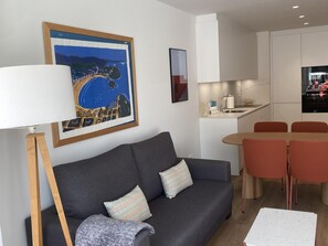 A modern living room with a grey sofa, two patterned pillows, a floor lamp, and a dining area with red chairs.