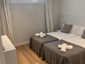 Une chambre d’hôtel avec deux lits, une literie grise, des rideaux blancs et un plancher en bois.