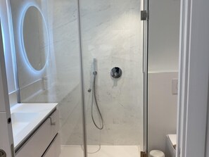 A modern bathroom with a glass-enclosed shower, a white marble wall, and a round mirror.