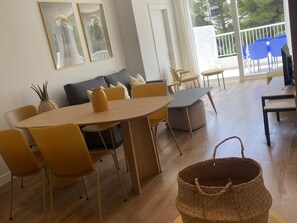 A modern living room with a round wooden table, yellow chairs, a grey sofa, and a large window with a view of blue chairs outside.