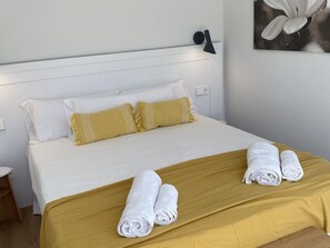 Ein ordentlich bezogenes Bett mit weißer und senffarbener Bettwäsche, zwei zusammengerollte Handtücher auf dem Bett und eine an der Wand befestigte Lampe.