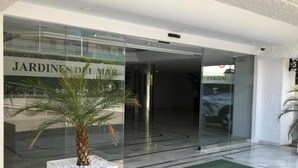 A modern building entrance with glass doors, a palm tree planter, and a doormat with 'Jardines del Mar' text.