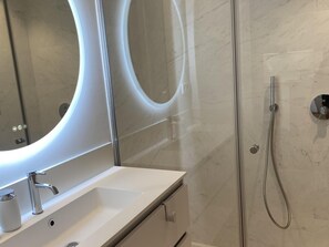 A modern bathroom with a white sink, a large mirror with a circular light, and a shower area.