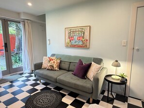 Un salon avec un sol à carreaux, un canapé gris agrémenté de coussins, une petite table d’appoint avec une lampe, et un tableau accroché au mur.