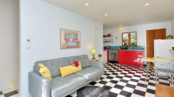 Una sala de estar con un sofá gris, piso a cuadros, una cocina con armarios rojos y un ventanal con vista a vegetación.