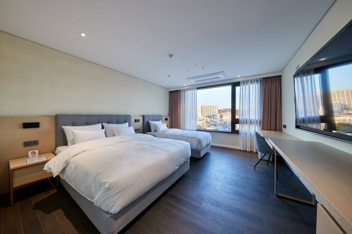 Ein Hotelzimmer mit zwei Betten, einem Schreibtisch und einem großen Fenster mit Blick auf die Stadt.