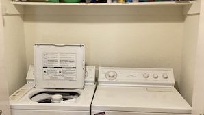 Une buanderie équipée d’un lave-linge et d’un sèche-linge, d’une étagère contenant des produits de nettoyage et d’une porte sur laquelle est apposée une étiquette d’avertissement.