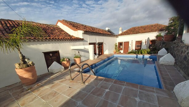 Una piscina con borde de baldosas, rodeada de edificios con techo de tejas, plantas en macetas y un muro de piedra.