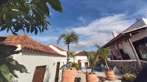 Una calle estrecha con arquitectura tradicional, incluyendo tejados de teja y paredes blancas, rodeada de macetas con plantas y un cielo azul despejado.