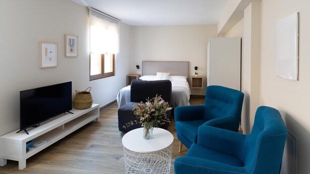 A modern, well-lit room with a bed, a TV, and two blue armchairs. - Housing León Azabachería 2A (León)