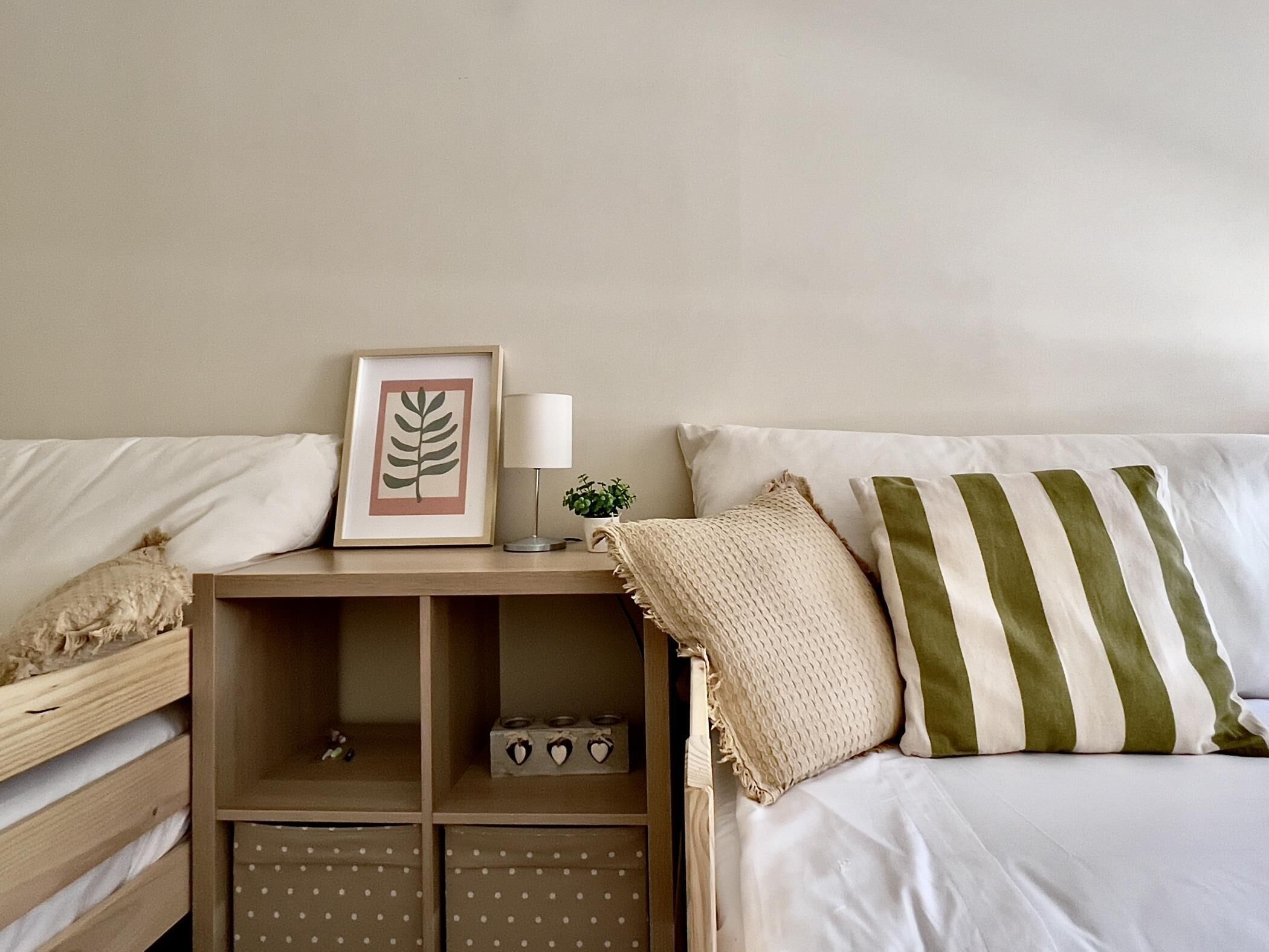 A neatly arranged bedroom with a bed, a bedside table with a framed leaf artwork, a lamp, and a small potted plant.