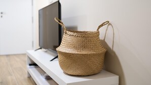 Una cesta tejida con asas sobre una consola blanca en una habitación con televisión y una puerta al fondo.