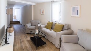 Una sala de estar moderna con un sofá, sillones y una mesa de centro. Hay un televisor sobre un mueble, una ventana con persianas y un cuadro en la pared.