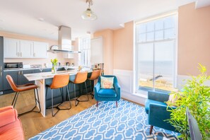 Une cuisine moderne avec des meubles blancs, un coin repas avec des chaises orange et une grande fenêtre donnant sur la plage.