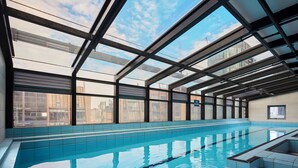 Ein großes Hallenbad mit klarem, blauem Wasser, umgeben von Glasdecke und -wänden, mit Blick auf die Stadtlandschaft.