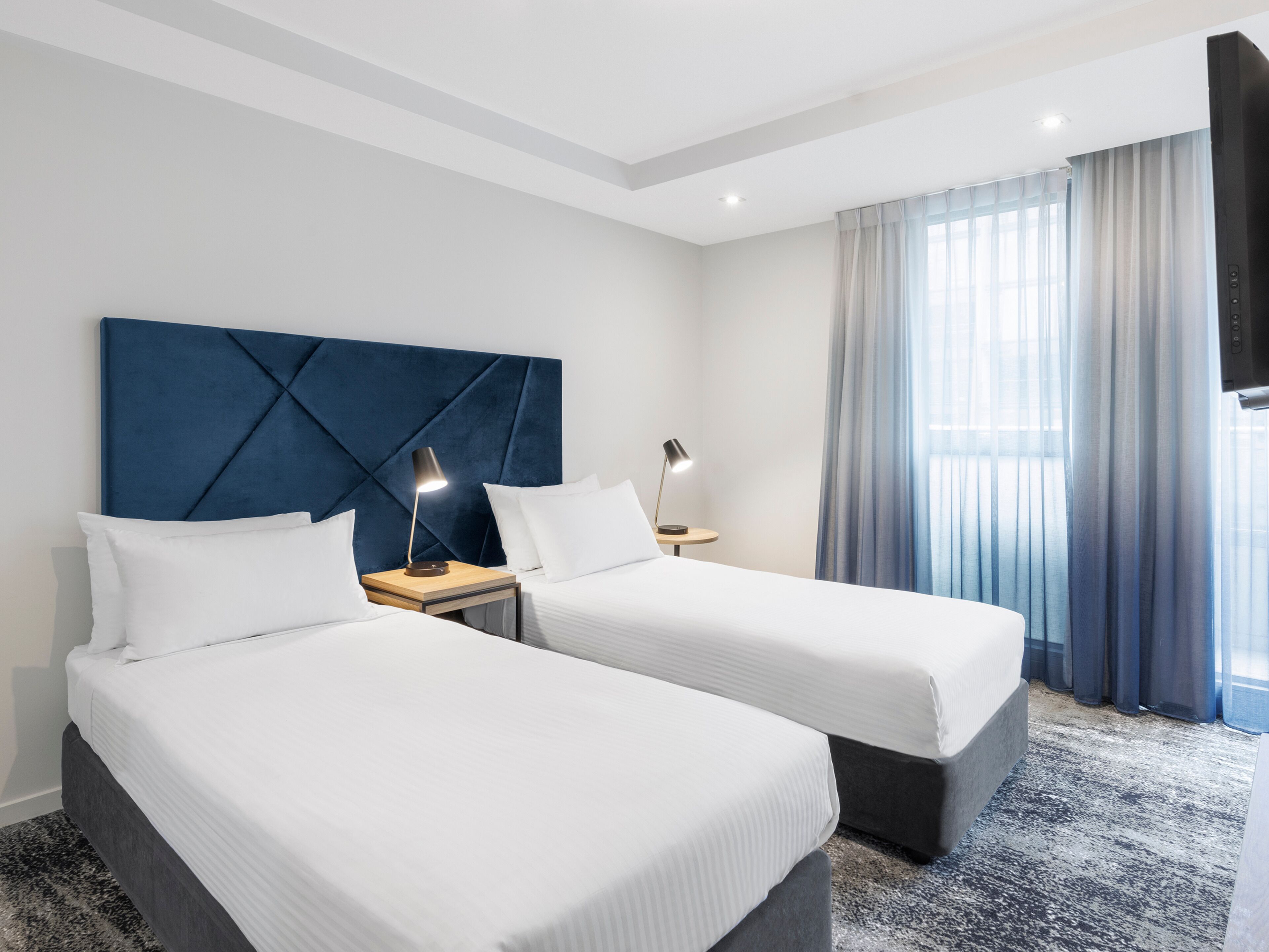 A hotel room with two beds, a large blue headboard, a TV, and a window with curtains.