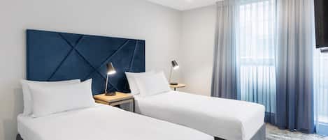 A hotel room with two beds, a large blue headboard, a TV, and a window with curtains.