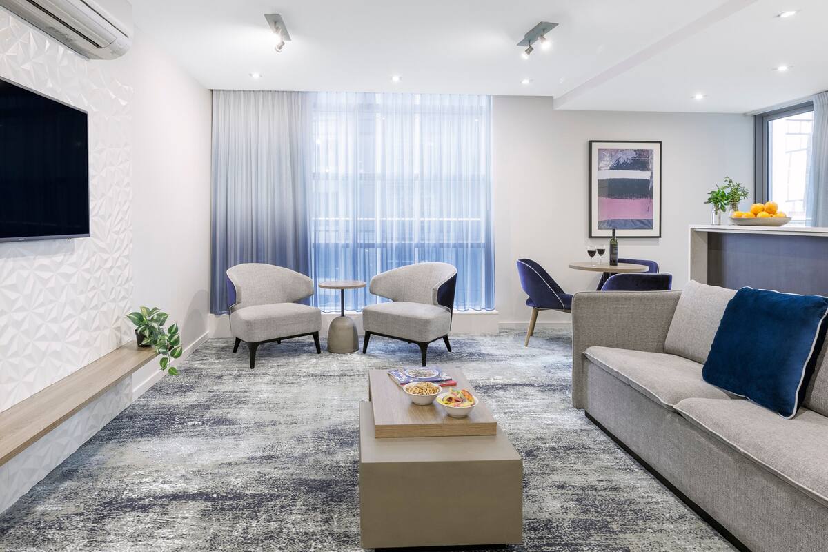 Una sala de estar moderna con un sofá, dos sillones, una mesa de centro con comida, un televisor y un comedor con mesa y sillas.