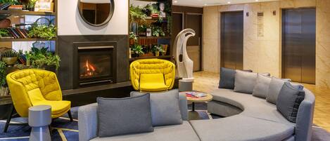 A modern lobby with a curved grey sofa, a yellow armchair, a fireplace, and a large mirror.