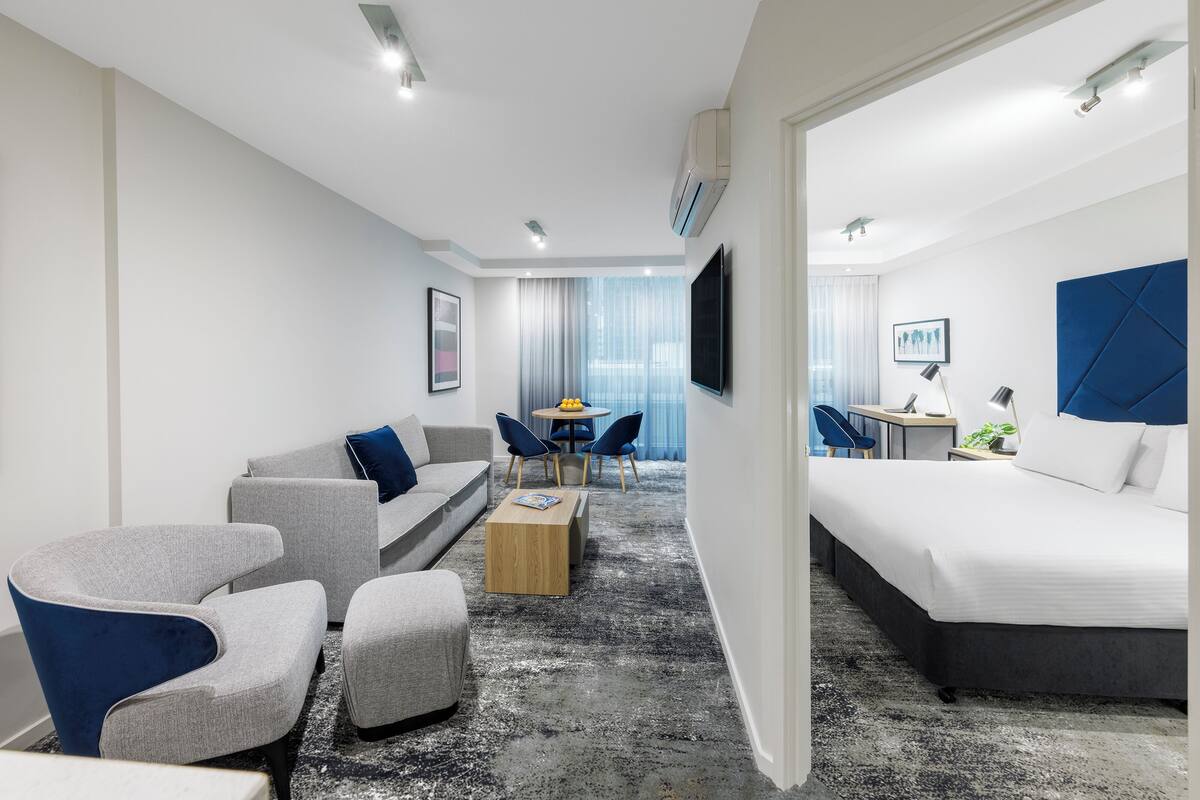 Habitación de hotel moderna con un sofá gris, una mesa de centro de madera, una cama con cabecera azul y un televisor montado en la pared.