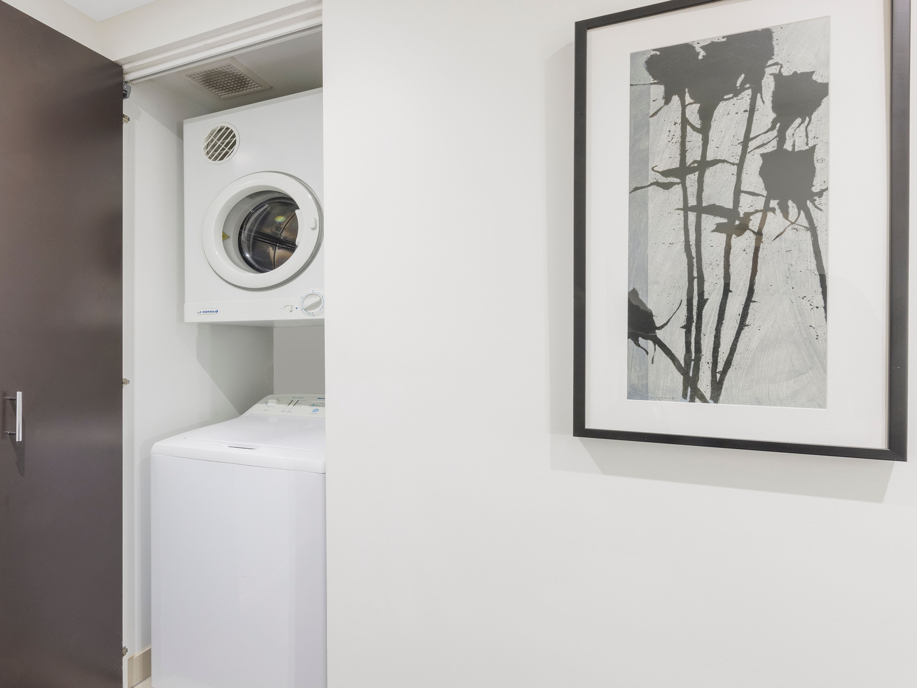 A laundry room with a washing machine and dryer, a framed abstract painting on the wall, and a door to the left.
