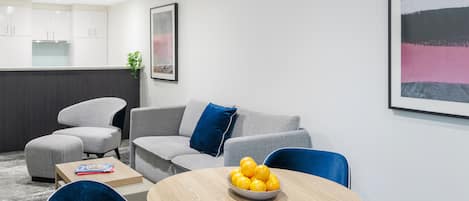 Una sala de estar moderna con un sofá gris, sillas azules, una mesa de centro de madera con un cuenco de naranjas, y una cocina al fondo.