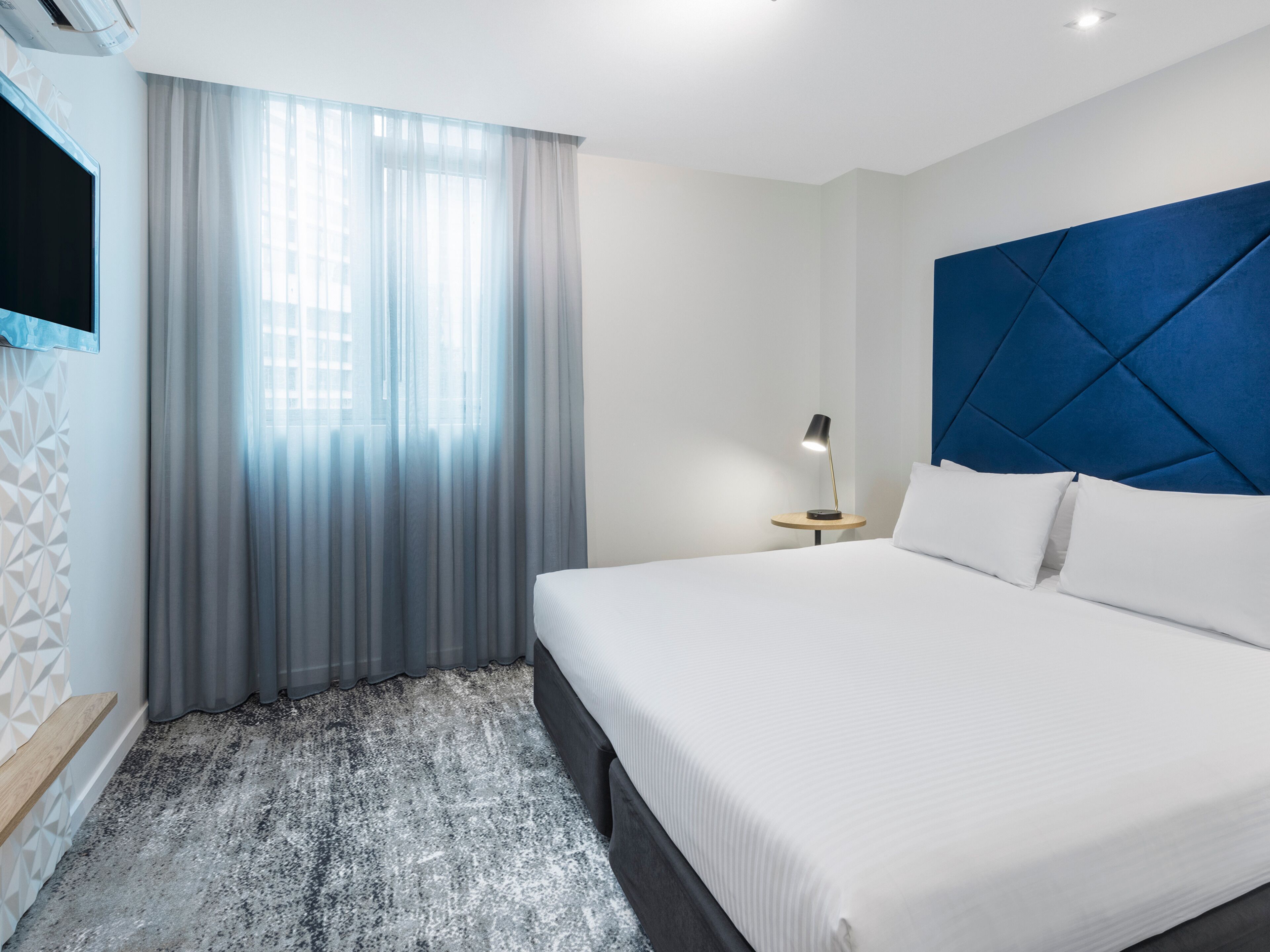 A modern hotel room with a large bed, a bedside table with a lamp, a TV mounted on the wall, and a window with sheer curtains.