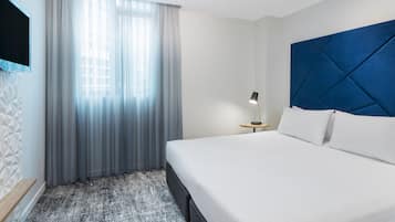 Et moderne hotelværelse med en stor seng, et sengebord med lampe, et tv ophængt på væggen og et vindue med lette gardiner.