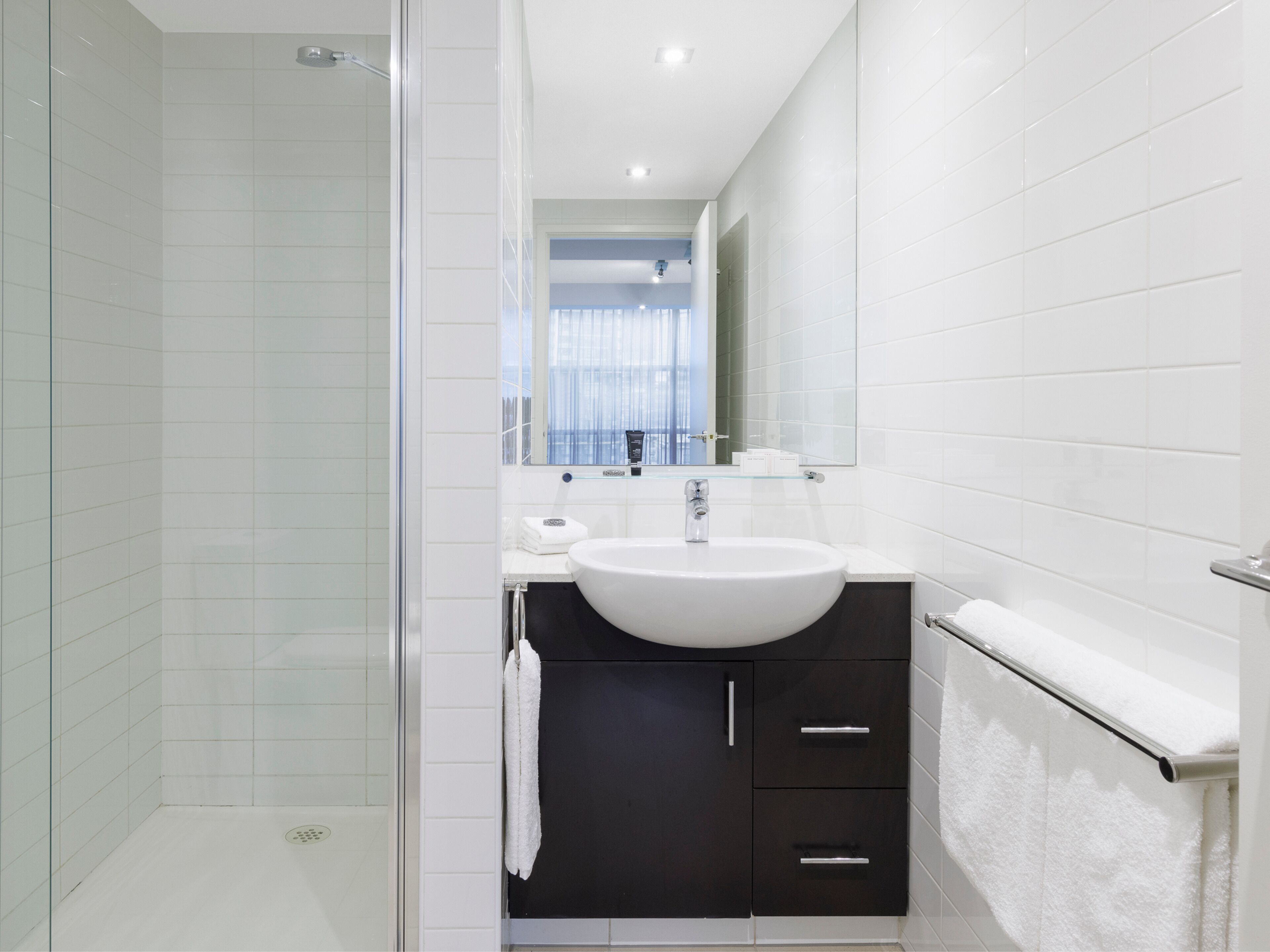 Een moderne badkamer met een glazen douchecabine, een witte wastafel, donkere kastjes en een spiegel boven de wastafel.