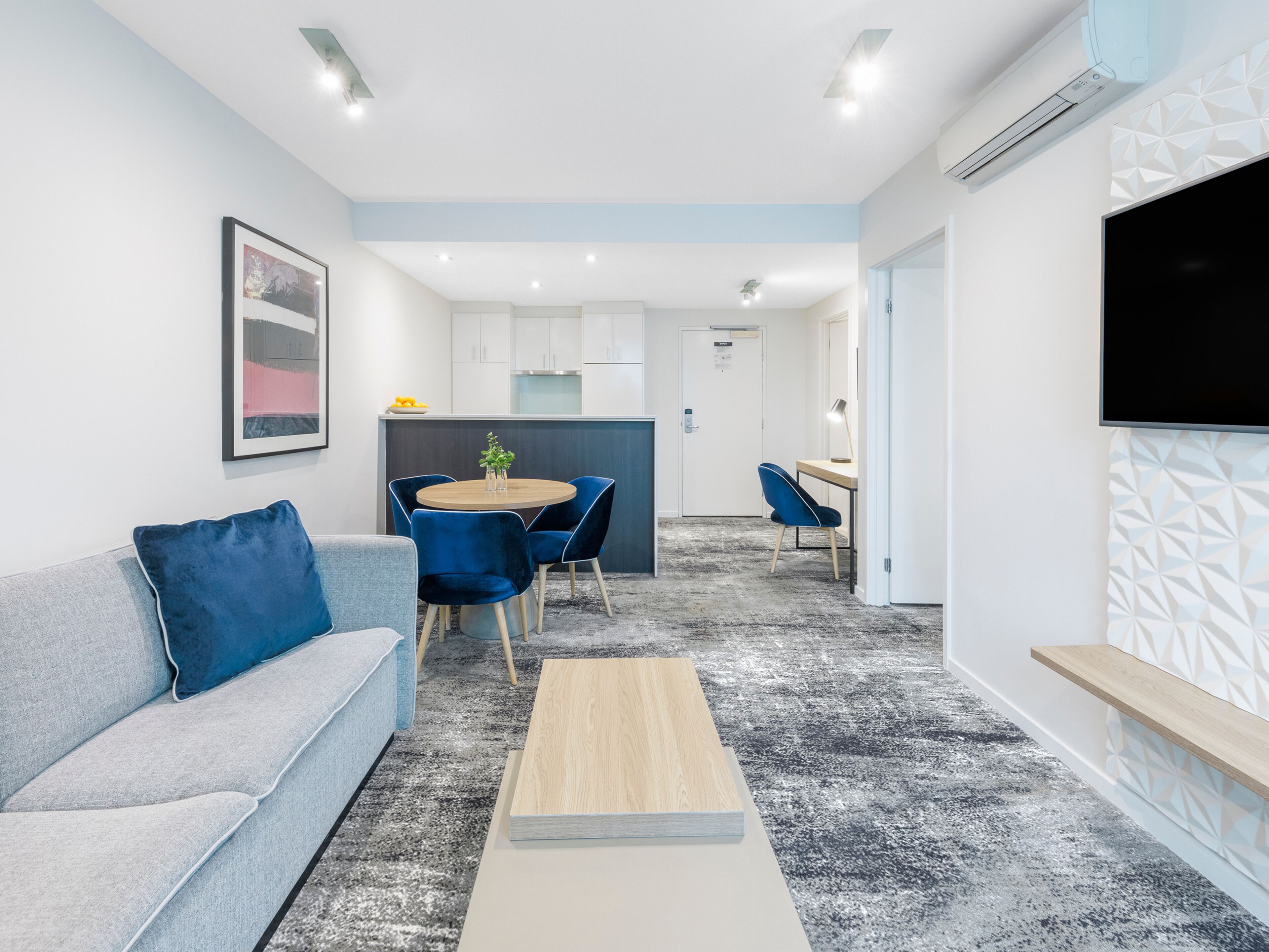 Un salon moderne avec un canapé gris, des fauteuils bleus, une table basse en bois et un téléviseur fixé au mur.