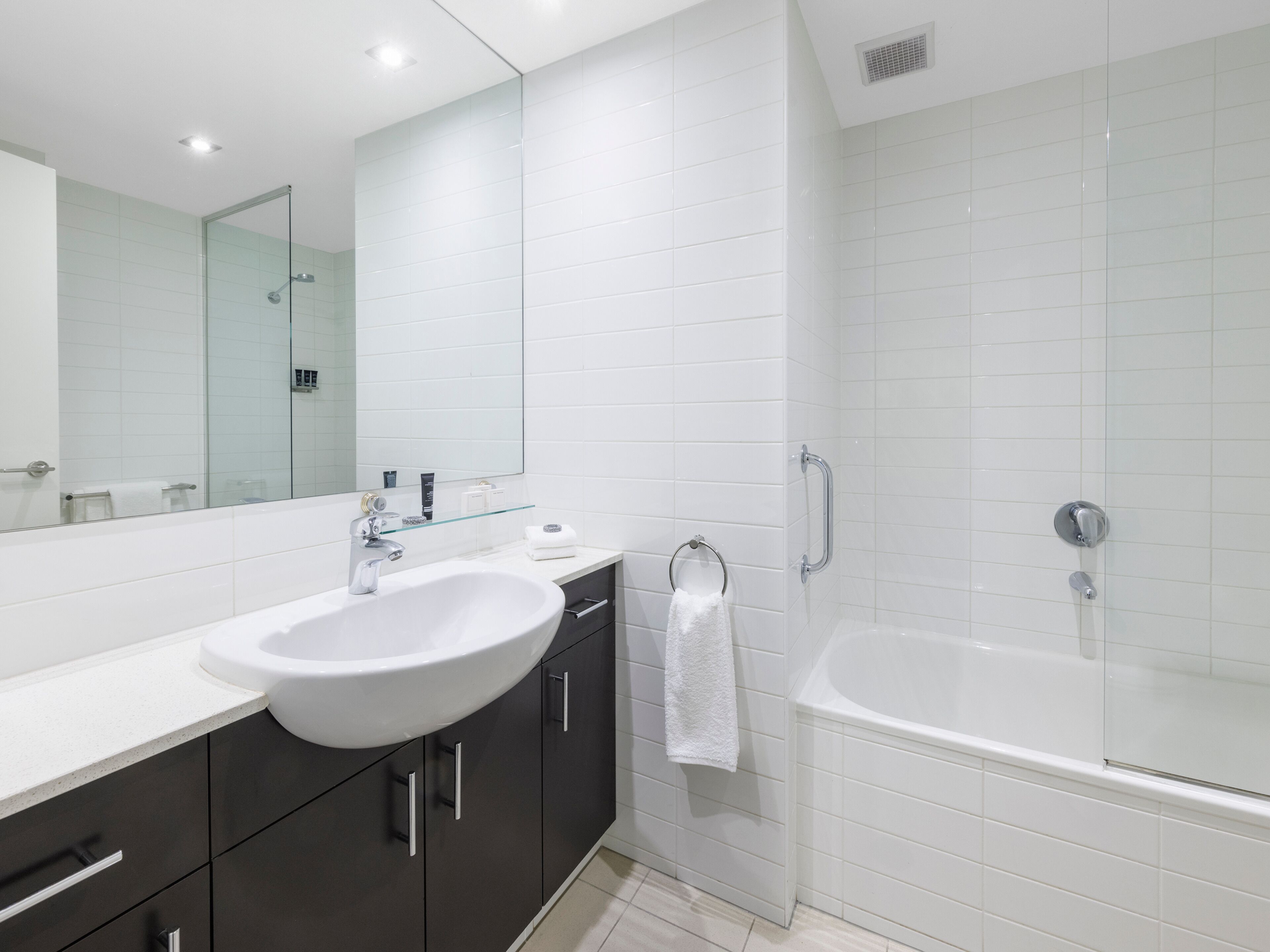 A modern bathroom with a white sink, dark cabinets, a glass shelf, and a white bathtub.