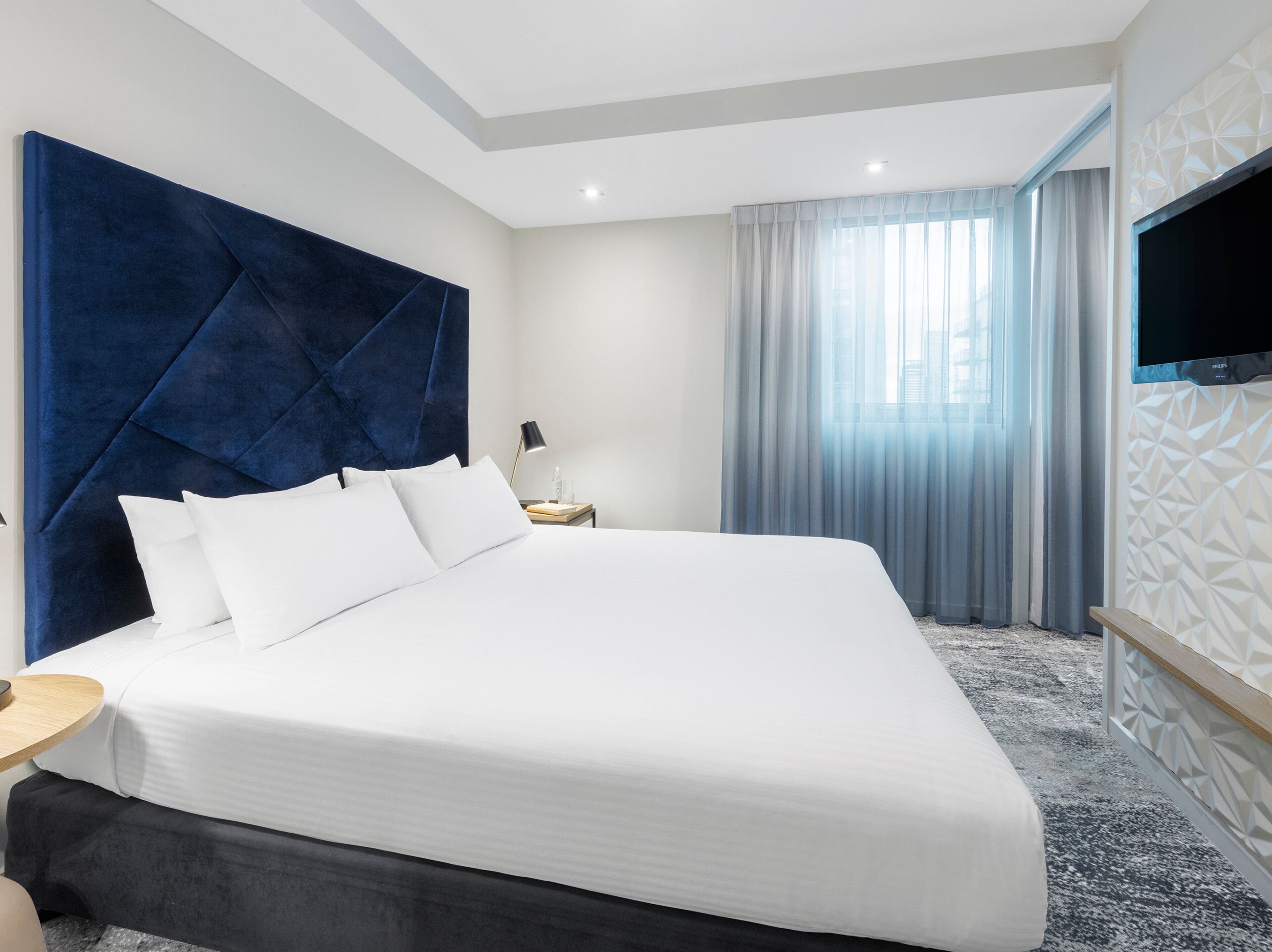 Una habitación de hotel moderna con una cama grande, un televisor de pantalla plana en la pared y una ventana con cortinas traslúcidas.