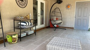 Ein Zimmer mit Fliesenboden, einem Korbsessel, einem Metallregal mit Dekorationsobjekten, einem Hängestuhl und einer Tür mit Sonnendekoration.