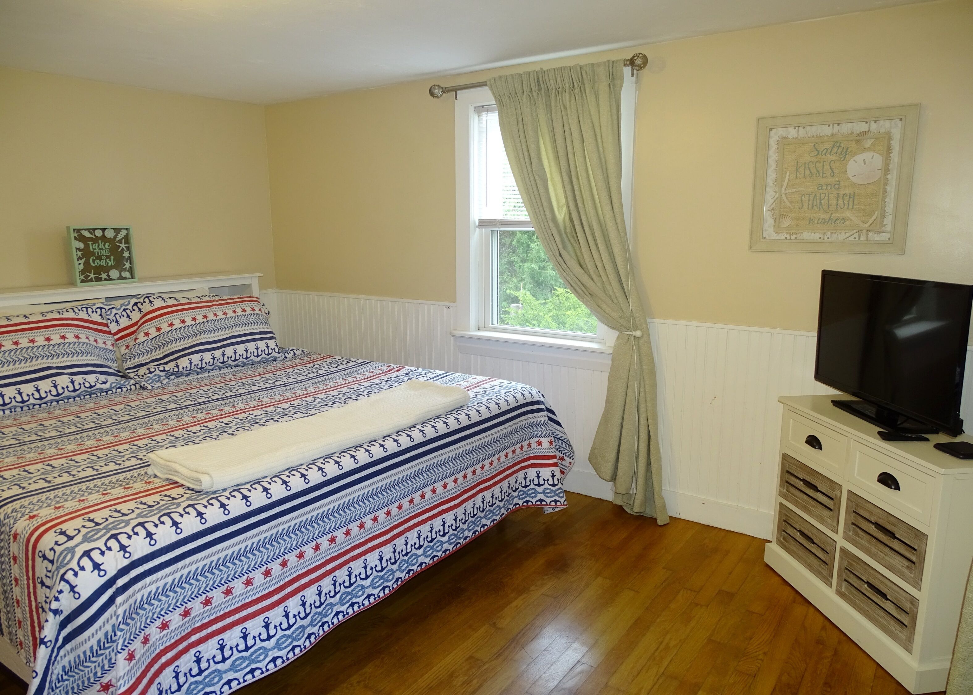 Un dormitorio con cama, televisión sobre un mueble, ventana con cortinas y un adorno en la pared.