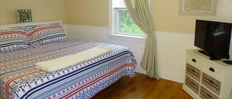 Un dormitorio con cama, televisión sobre un mueble, ventana con cortinas y un adorno en la pared.