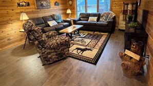 Un interior acogedor de cabaña de madera con dos sofás, un sillón estampado, una mesa de centro y un letrero de bienvenida en un aparador de madera.