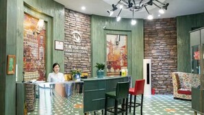 Un área de recepción con una recepcionista, un mostrador, una pared verde con diseño de piedra, un suelo estampado y una lámpara en el techo.